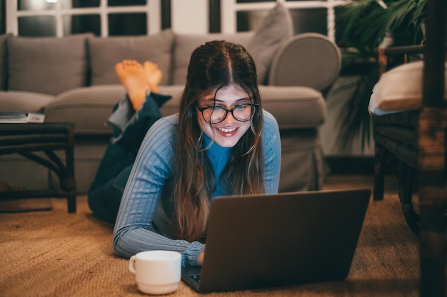 Een aantrekkelijke vrolijke en jonge vrouw met een bril die 's nachts computer op het tapijt thuis gebruikt