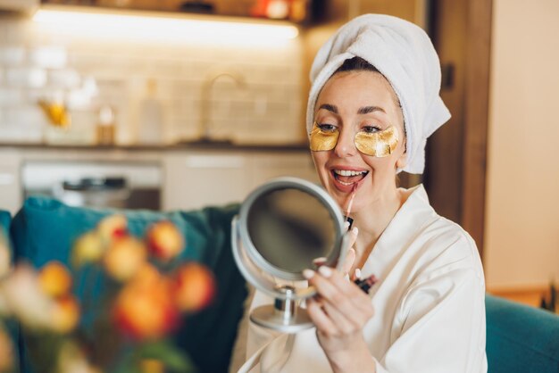 Een aantrekkelijke vrouw met huidverzorging onder de ogen pleisters die lipgloss aanbrengt terwijl ze van de ochtend thuis geniet.