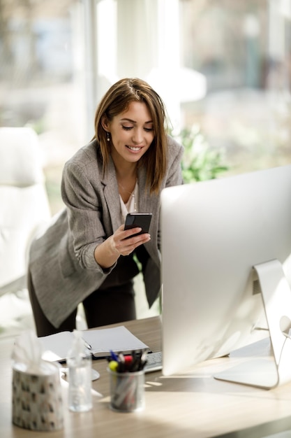 Een aantrekkelijke zakenvrouw die smartphone gebruikt terwijl ze op de computer in haar kantoor werkt.