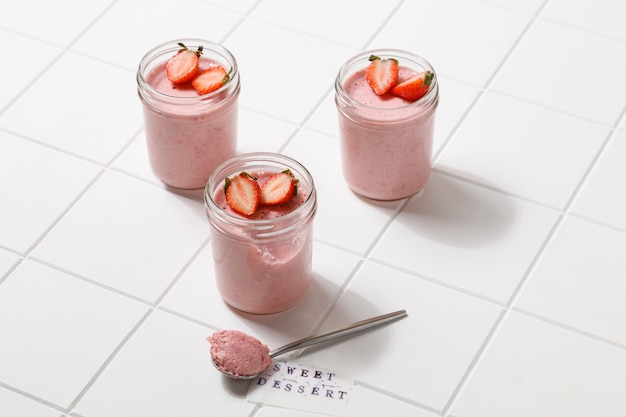 Een aardbeiendessertmousse in glas met lepel op wit bord op witte tegelachtergrond