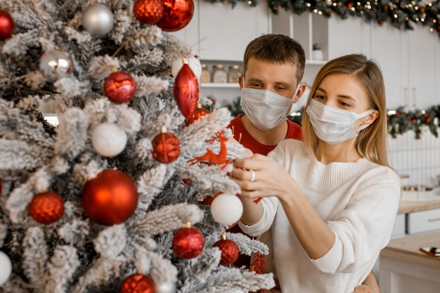 Een aardig, aantrekkelijk stel dat een gezichtsbeschermend medisch masker draagt, concentreert zich op het versieren van de kerstboom thuis