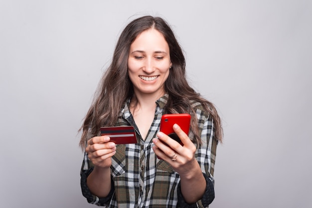 Een aardige jonge vrouw houdt een telefoon vast en met een andere hand een creditcard bij een grijze muur