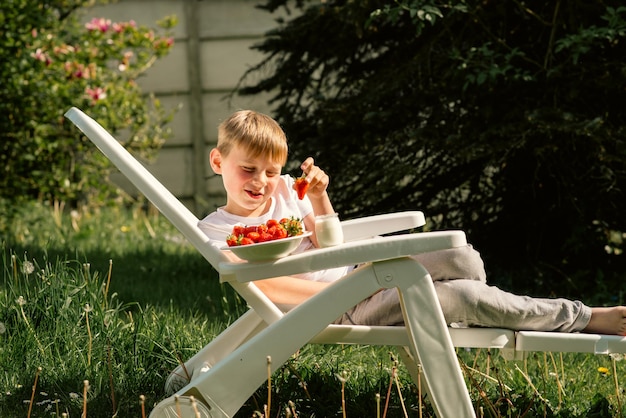 Een achtjarige jongen eet in de zomer aardbeien met yoghurt in de tuin