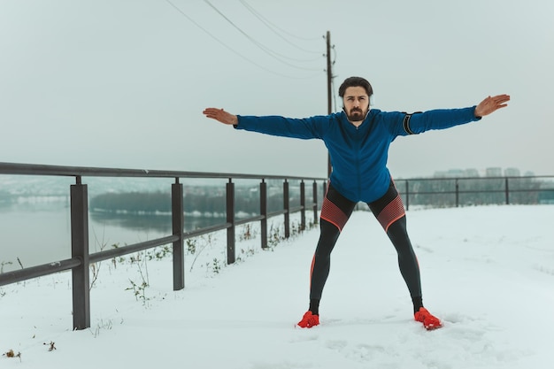 Een actieve jongeman, met koptelefoon op zijn oren, rekt zich uit en doet oefeningen in de openbare ruimte tijdens de wintertraining buiten.