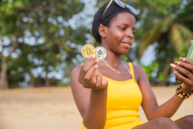 Een Afrikaanse handelaar met Bitcoin en etherische munten op het strand controleert de markt met haar telefoon