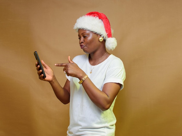 Foto een afrikaanse vrouw die met een vinger wijst en naar de smartphone in haar hand kijkt met een kerstmuts op haar hoofd