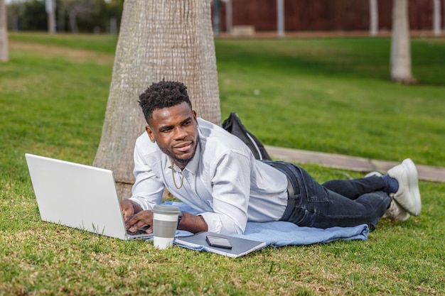 Een Afro-Amerikaanse man die aan zijn laptop werkt en op de grond ligt in het park.