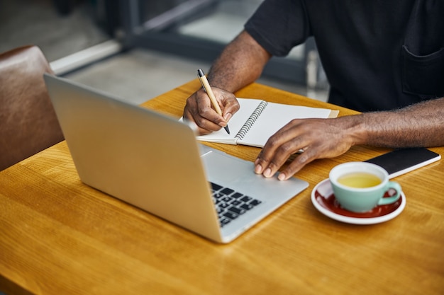 een Afro-Amerikaanse man zit achter een laptop met koffie en smartphone en schrijft in een notitieboekje