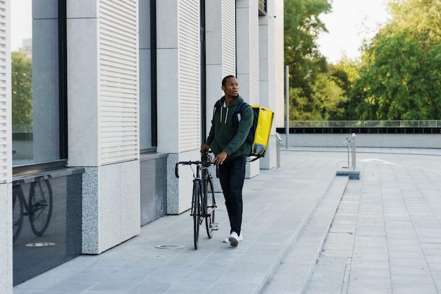 Een Afro-Amerikaanse mannelijke koerier met een rugzak loopt naast een fiets en zoekt de ingang van het gebouw