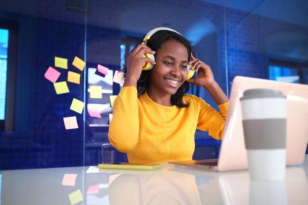 Een afro-amerikaanse vrouw zet een draadloze headset op terwijl ze op haar laptop op kantoor werkt