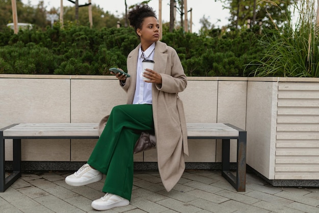 Een Afro-Amerikaanse vrouw zit op een bankje in het park met een laptop en een mobiele telefoon eruit