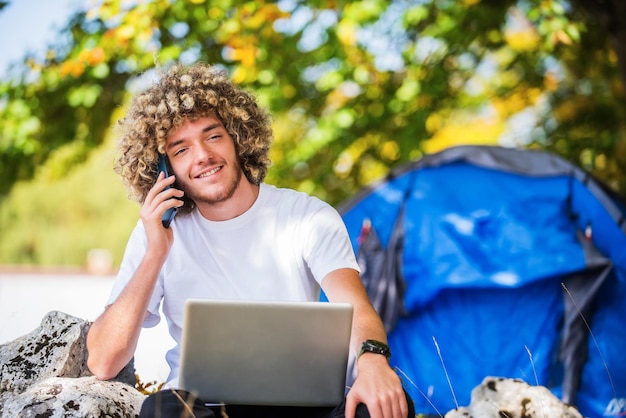 Een Afro-man zit in de natuur en gebruikt een smartphone en een laptop voor een online vergadering