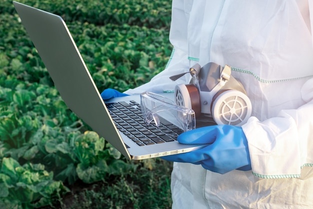 Een agronoom gekleed in een wit beschermend pak, masker en blauwe handschoenen houdt laptop tegen het veld met oogst. Problemen van het concept van chemicaliën en pesticiden.