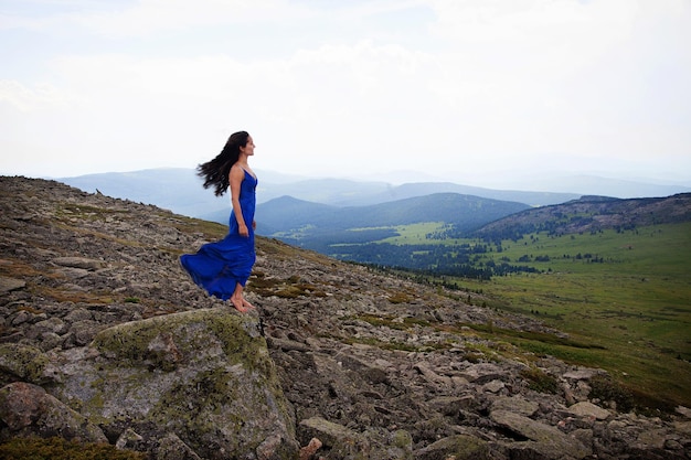 Een altai-meisje in een blauwe jurk staat bovenop de sarlyk-berg boven een klif tussen rotsen en stenen. toerisme concept.