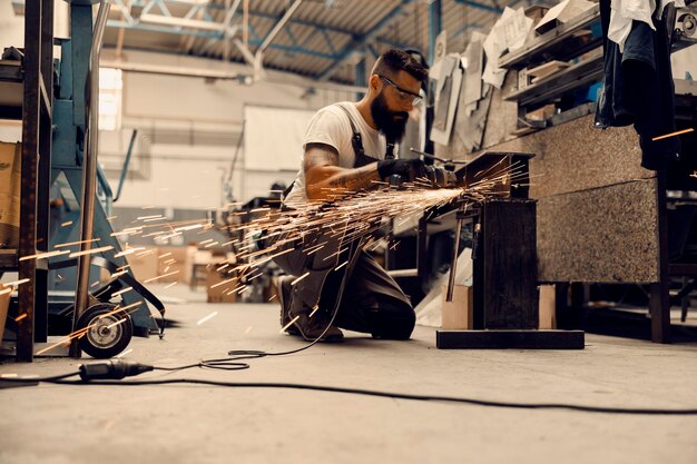 Een arbeider in de zware industrie knielt in een faciliteit en verwerkt metalen onderdelen met een slijpmachine