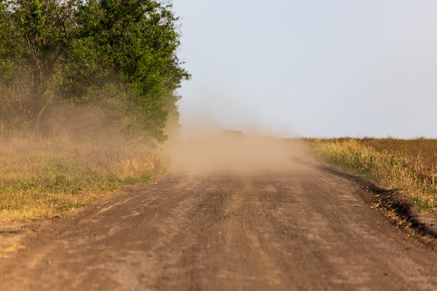 Een auto in een stofwolk die in de verte wegrijdt langs een landelijke weg tussen een veld en bomen