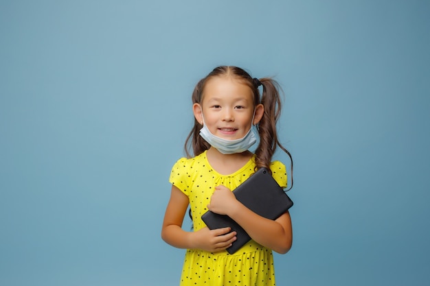 Een Aziatisch meisje met een masker op haar gezicht houdt een tablet in haar handen in de Studio op een blauw