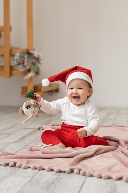 Een Aziatische baby van 9 maanden in een kerstkostuum speelt met een nieuwjaarsspeelgoed op een deken die op de grond ligt en kijkt in de camera met haar ogen wijd open en lacht