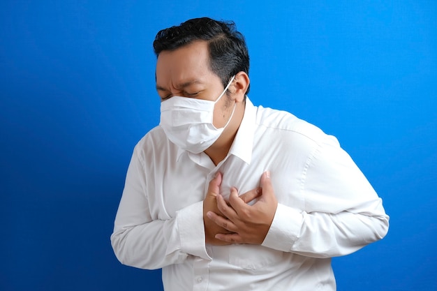 Een aziatische dikke man met een masker die pijn in zijn borst voelt, gebaar met linkerborst. Blauwe achtergrond