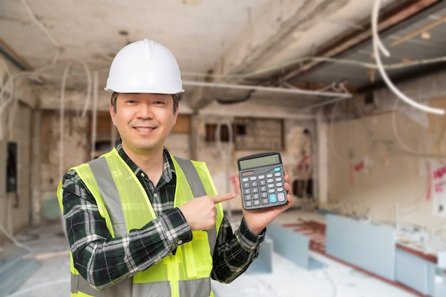 Een Aziatische man op middelbare leeftijd die een witte het werk GLB en een werkpak draagt dat een calculator in zijn hand houdt.