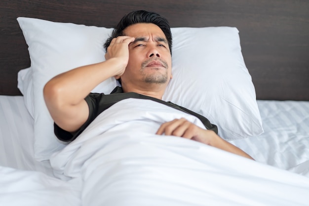 Een Aziatische man slaapt met hoofdpijn en houdt zijn hoofd angstig vast in het witte bed in de slaapkamer.