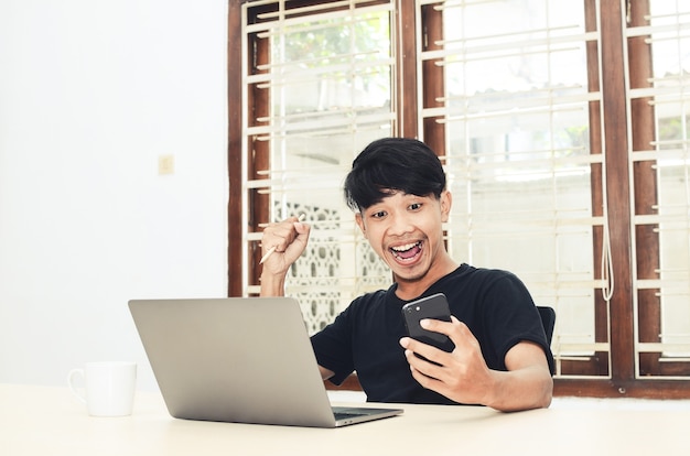 Een Aziatische man zit voor de laptop met een vrolijke uitdrukking terwijl hij naar de telefoon kijkt