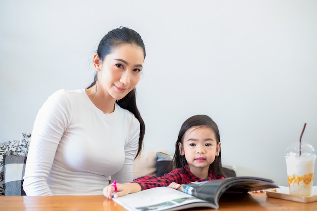 Een Aziatische moeder leert haar dochter om een boek te lezen tijdens de pauze van het semester op de levende tafel en met koude melk op tafel thuis. Educatieve concepten en activiteiten van het gezin