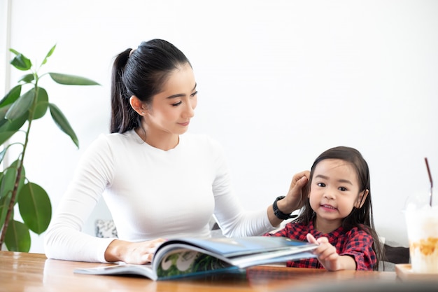 Een Aziatische moeder leert haar dochter om een boek te lezen tijdens de pauze van het semester op de levende tafel en met koude melk op tafel thuis. Educatieve concepten en activiteiten van het gezin