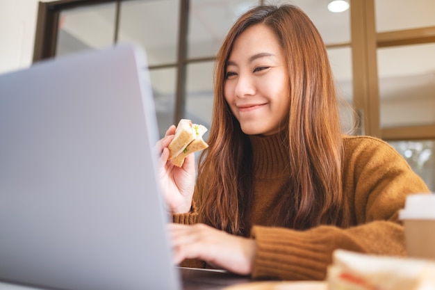 Een aziatische vrouw die een volkoren broodje vasthoudt en eet terwijl ze op een laptop werkt