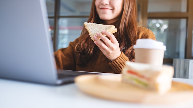 Een aziatische vrouw die een volkoren broodje vasthoudt en eet terwijl ze op een laptop werkt