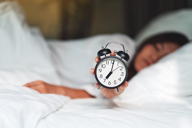 Een Aziatische vrouw die en een wekker houdt toont terwijl het slapen op een wit comfortabel bed in de ochtend