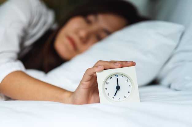 Een Aziatische vrouw die 's ochtends een wekker uitzet terwijl ze op een wit, knus bed slaapt