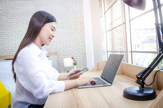 Een Aziatische vrouw die thuis zit Houd de smartphone vast en typ de informatie op de laptop