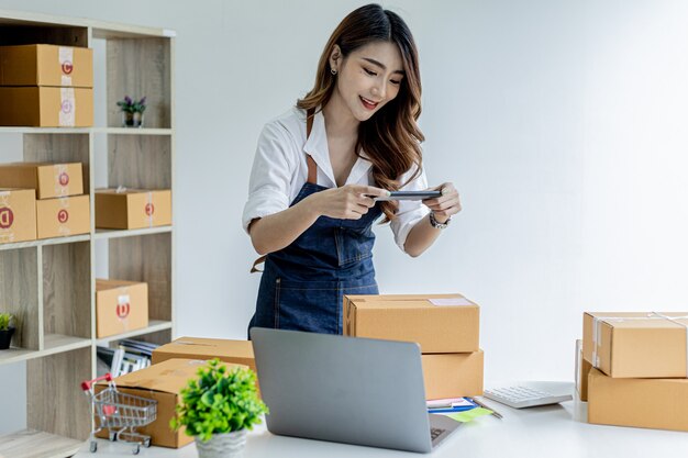 Een Aziatische vrouw gebruikt een smartphone om foto's te maken voor een pakketdoos als bewijsmateriaal, ze heeft een online winkel, ze verpakt en verzendt via een particuliere rederij. Online verkoopconcept.