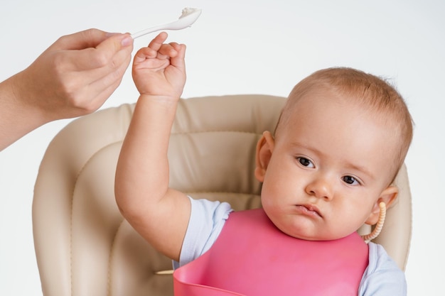 Een baby die op een stoel zit, duwt de hand van zijn moeder weg met een lepel met eten