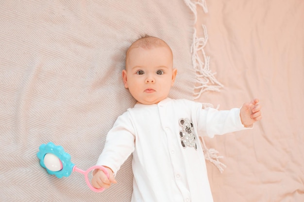 Een baby in een witte jumpsuit ligt thuis op een bed met een rammelaar