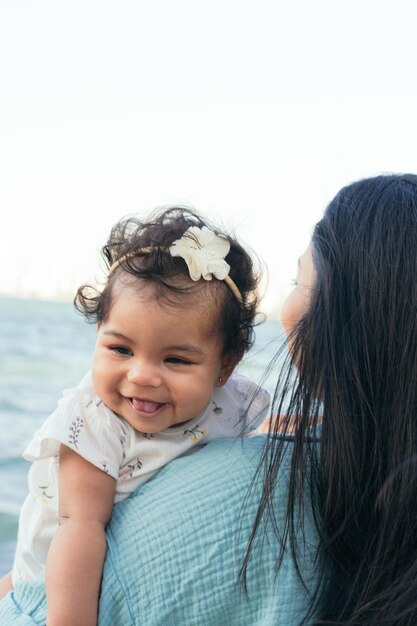 Een baby lacht terwijl haar moeder haar in haar armen houdt. Op de achtergrond zijn de golven van de zee te zien