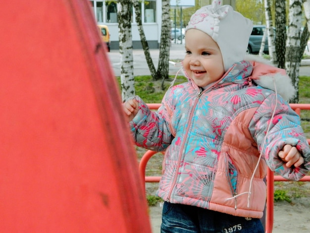 Een baby staat voor een rode paal en het meisje draagt een roze jasje met de letters j.