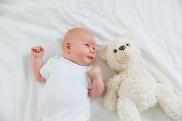 Een baby van 2 maanden in een witte bodysuit ligt thuis op een wit bed naast een teddybeer-uitzicht van bovenaf pasgeboren