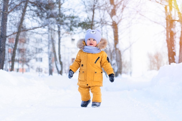 Een baby ziet voor het eerst sneeuw. Portret van een peuter van 15-20 maanden oud in gele warme kleren in een winterpark die vrolijk naar sneeuwvlokken kijkt