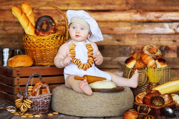 Een babybakkersjongen in een koksmuts en schort met een bos bagels om zijn nek bereidt deeg voor op het bakken tegen de achtergrond van bakkerijproducten.