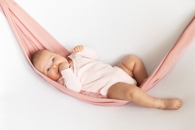 Een babymeisje in een roze bodysuit ligt in een roze hangmat op een witte neutrale achtergrond en zuigt aan haar vinger.