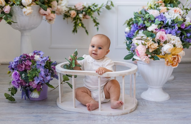 Een babymeisje in een wit rompertje met kant zit in een stoel op een achtergrond met bloemen