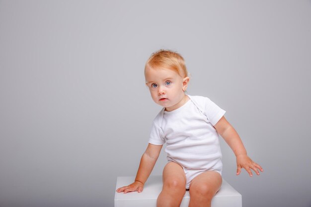 een babymeisje in een witte bodysuit zit op een witte kubus op een witte achtergrond