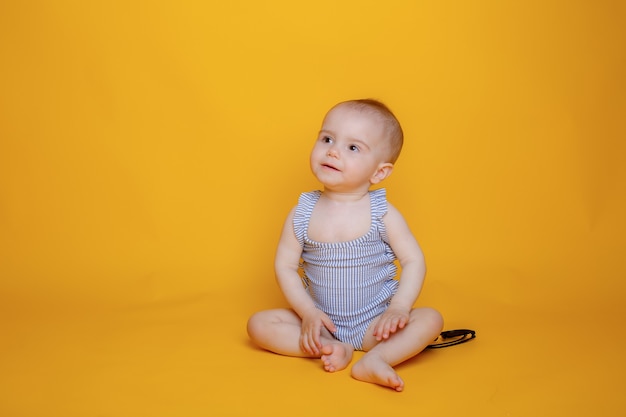 Een babymeisje in een zwempak met een hoed zit op een gele achtergrond