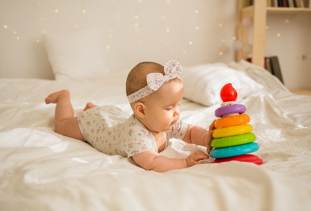 Een babymeisje ligt en speelt met een piramide op een witte deken op bed