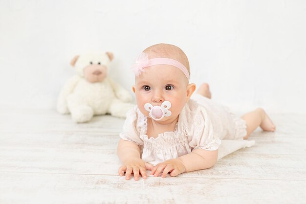 Een babymeisje met een fopspeen in haar mond in een witte jurk met een speeltje ligt zes maanden op haar buik