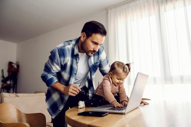 Een babymeisje speelt thuis met een laptop terwijl haar vaderondernemer naar het rapport kijkt