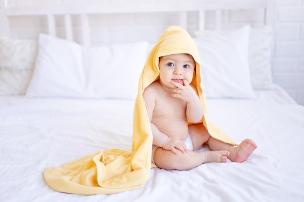 Een babymeisje van zes maanden op een bed met een gele handdoek op haar hoofd na het baden of wassen van een klein kind op een katoenen bed thuis het concept van zorg en hygiëne