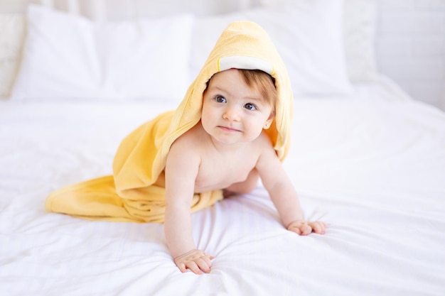 Een babymeisje van zes maanden op een bed met een gele handdoek op haar hoofd na het baden of wassen van een klein kind op een katoenen bed thuis het concept van zorg en hygiëne